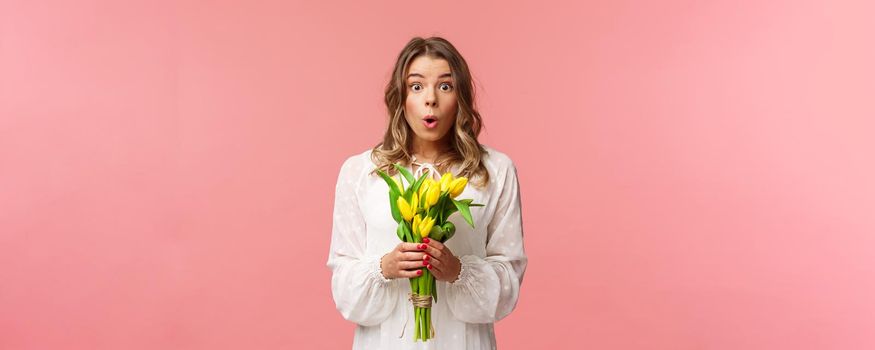 Holidays, beauty and spring concept. Portrait of surprised and amazed blond girl in white dress, holding yellow tulips, receive flowers being amused and happy, standing pink background.