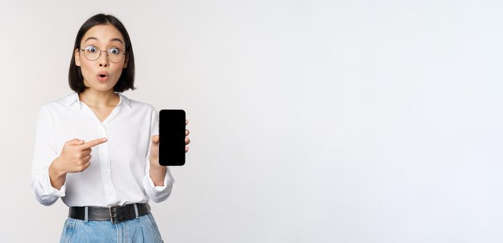 Enthusiastic young asian woman pointing finger at smartphone screen, showing advertisement on mobile phone, white background.