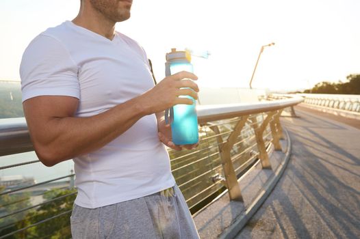 Cropped view of a muscular build Caucasian man, fit athlete with beautiful aesthetic body holding a bottle with water for rehydration and refreshment after heavy outdoor workout at dawn