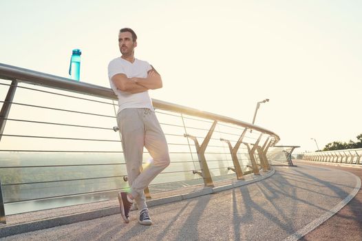 Attractive middle aged muscular build European man, fit athlete with beautiful aesthetic body relaxing after heavy outdoor workout at dawn, standing on a glass city bridge