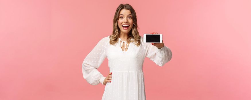 Portrait of cheerful, upbeat attractive blond caucasian girl in white dress, showing smartphone display, hold mobile phone horizontal smiling camera amazed, stand pink background.