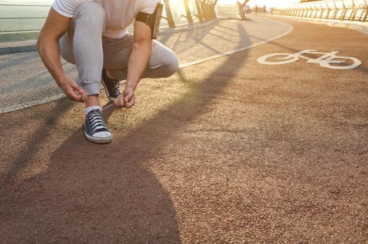 Close-up. Athlete runner tying shoelaces, ready for running. Sunbeams falling on a city bridge treadmill. Cardio workout, outdoor sports training, Motion, endurance and healthy lifestyle concept