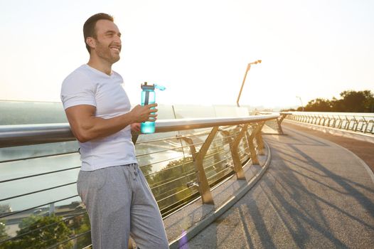 Happy middle aged fit European man, muscular build sportsman athlete drinking water while relaxing after morning jog on the city bridge. Rehydration, healthy active lifestyle concept