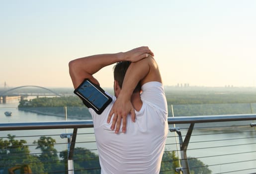 Rear view of an athlete doing sports. Muscular build sportsman standing on a city bridge and stretching his arms, warming up his body during a morning workout