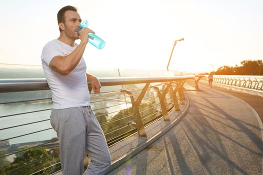 Waist length portrait of handsome attractive middle aged European muscular build man, athlete drinking water after morning jog on the city bridge treadmill at dawn. Sport, endurance, healthy lifestyle