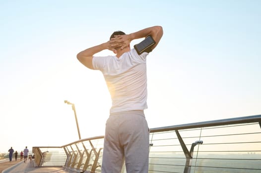 Keep you body fit. Rear view of a muscular sportsman in a white t-shirt, wearing a smartphone holder, doing a warm-up at dawn, enjoying a morning workout before jogging