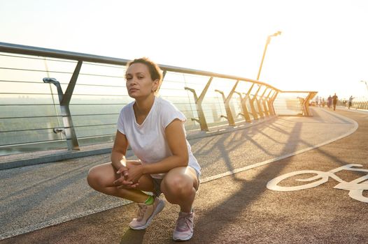 Portrait of charming middle aged beautiful woman in white t-shirt and blue denim shorts squatting on treadmill and looking at camera while enjoying early morning walk at sunrise