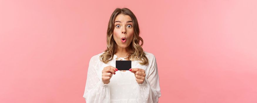 Close-up portrait of excited and amused young girl describe new features of her bank, received new credit card, say wow, folding lips thrilled and amazed, standing pink background.