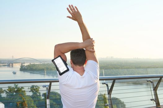 Athlete goes in for sports in an urban environment. Rear view of an athlete exercising on the city bridge, warming up and stretching the body during a morning workout outdoors at dawn