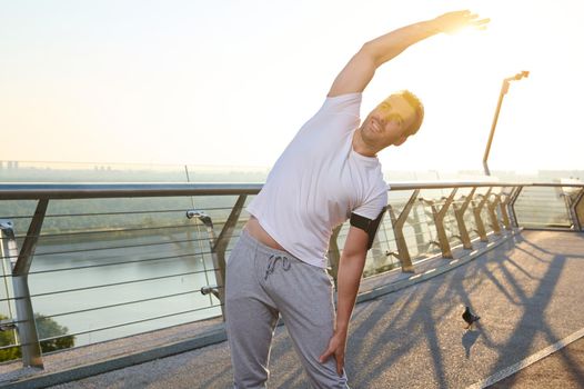 Middle aged Caucasian athlete sportsman stretches his arm and shoulder outdoors, doing warm-up, enjoying an early morning workout at dawn, standing on a city bridge and admiring the beautiful nature.