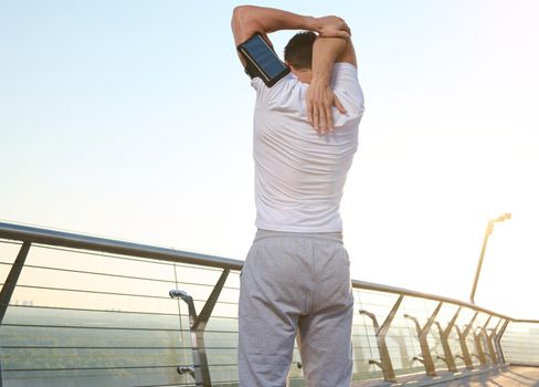 Middle aged muscular athlete stretches his arm and shoulder outdoors and does a warm-up, enjoying a morning workout on a summer sunny day, standing on a city bridge and admiring the beautiful nature