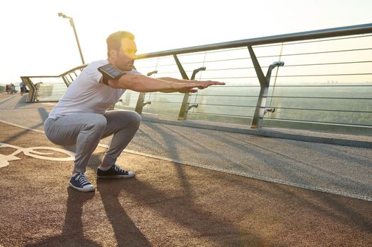 Middle-aged athlete, sportsman, muscular build European man is engaged in sports on a city bridge at dawn, performs squats, kneads muscles of the body. The concept of a healthy active lifestyle