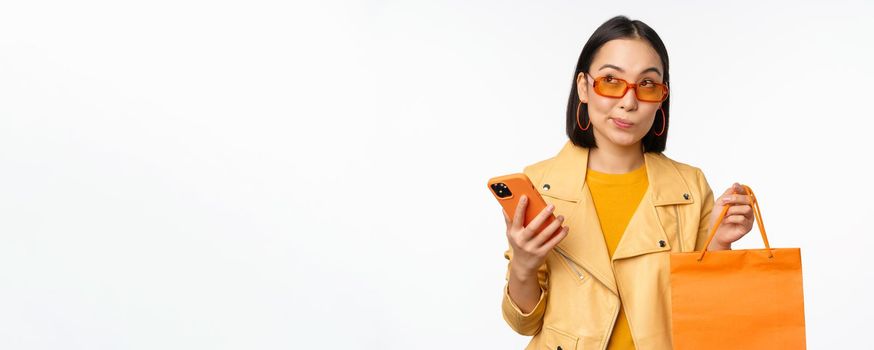 Stylish asian girl shopper, wears sunglasses, holding shopping bag and smartphone, going for discounts in stores, standing over white background.