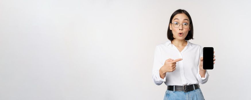 Enthusiastic young asian woman pointing finger at smartphone screen, showing advertisement on mobile phone, white background.