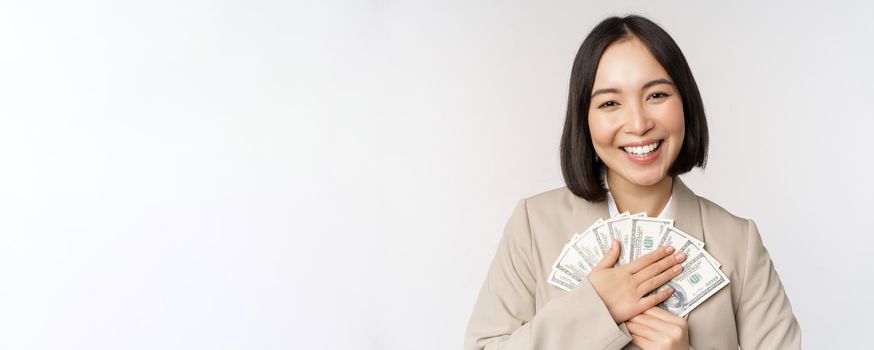 Happy asian businesswoman holding cash, hugging dollars money and smiling, standing over white background in suit.