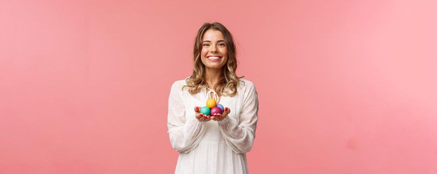 Holidays, spring and party concept. Portrait of tender, lovely blonde girl in white dress, holding Easter painted eggs, celebrating orthodox day, smiling cheerful share positivity, pink background.