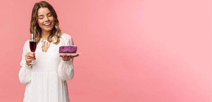 Holidays, spring and party concept. Portrait of tender, elegant young blond woman holding glass of wine and cake with lit candle, smiling pleased, celebrating birthday, pink background.