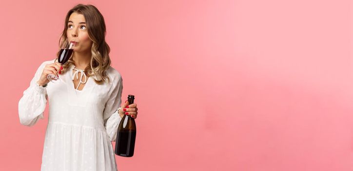 Holidays, spring and party concept. Portrait of carefree independent cute blond woman sipping wine from glass, holding bottle and tasting drink, look up, celebrating with friends, pink background.
