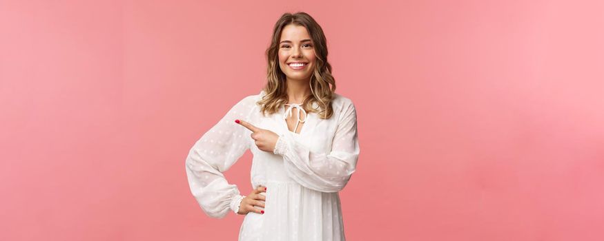 Pleasant good-looking smiling blond woman with blond short wavy hair, white tender dress, looking camera and pointing finger left to show you best price, good offer, promo concept, pink background.