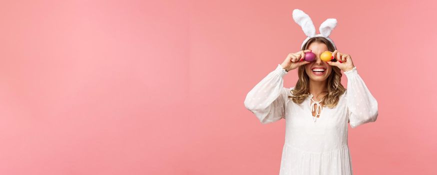 Holidays, spring and party concept. Portrait of lovely, tender smiling woman in rabbit ears and white dress celebrating Easter day, holding painted eggs on eyes and grinning, pink background.