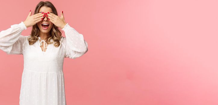 Excited cute blond girl in white spring dress waiting for gifts at surprise party, cover eyes blindfolded standing pink background, open mouth in amazement and anticipation.