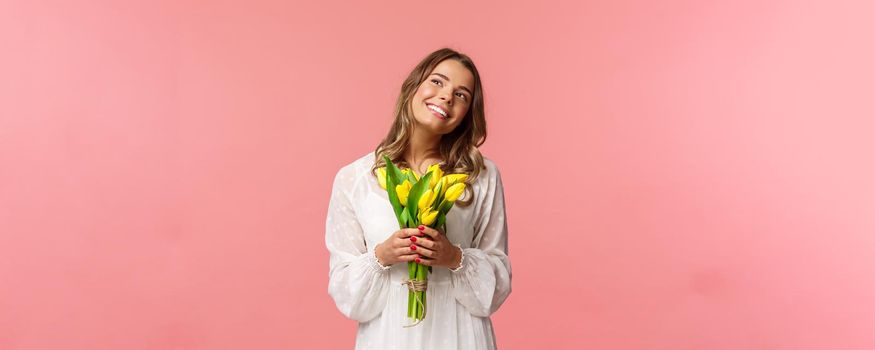 Holidays, beauty and spring concept. Portrait of dreamy, happy blond girl feeling romantic daydreaming about her girlfriend, holding yellow tulips, wear white dress, standing pink background.