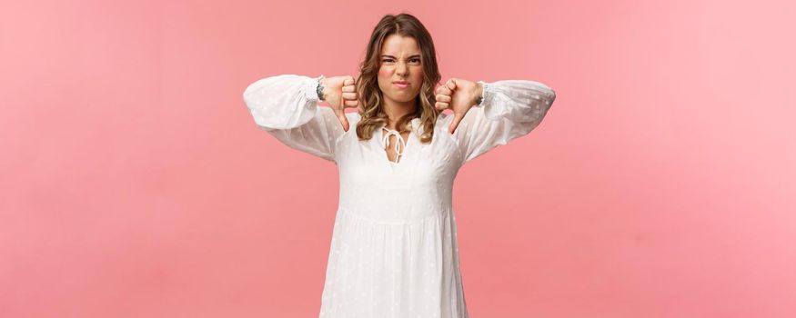 Portrait of picky young blond caucasian woman in white dress over pink background expressing dislike, show thumbs-down and grimacing in aversion, disappointed with bad quality.