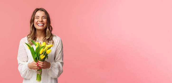 Holidays, beauty and spring concept. Portrait of happy excited charming blond girl receive flowers, buying yellow tulips herself, smiling and laughing joyfully, stand pink background.