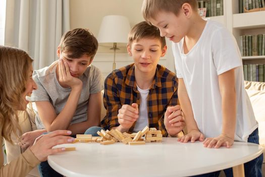 Three boys and a woman in the room are playing a board game made of wooden rectangular blocks, vivid emotions from the collapsed tower.