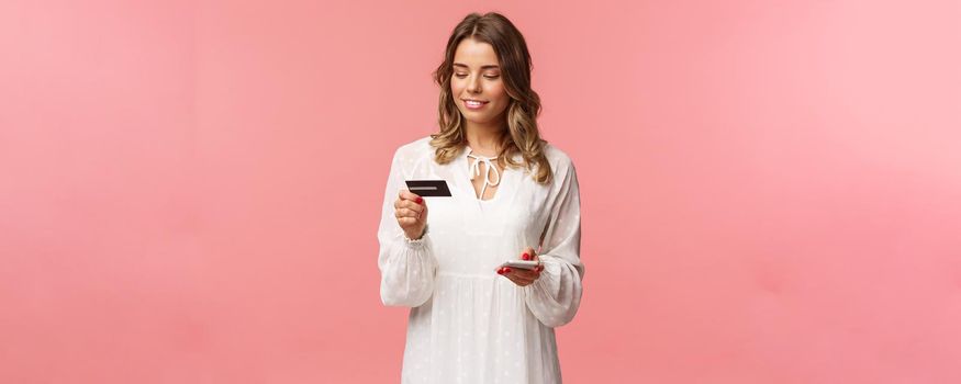 Portrait of attractive and cute blond girl with short curly hairstyle, white dress, holding smartphone and credit card, insert digit numbers to register in shopping app, buy online, pink background.