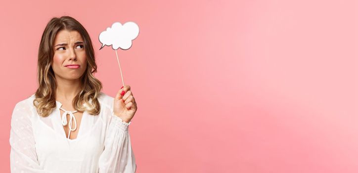 Spring, happiness and celebration concept. Close-up of skeptical and doubtful good-looking blond girl holding commend cloud near head as if having thought, grimacing and squinting from dislike.