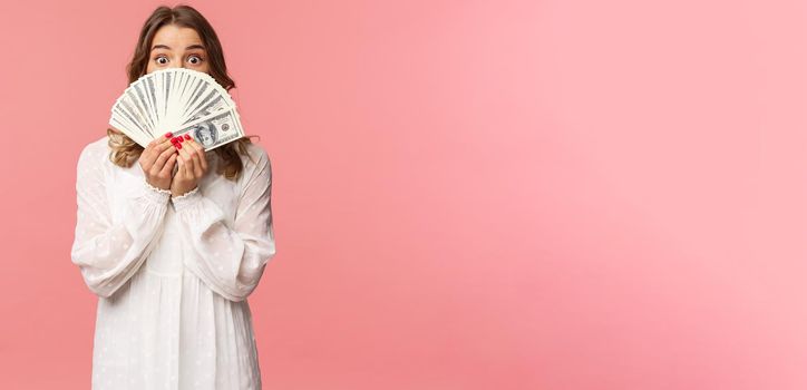 Portrait of amazed and excited cute feminine blond girl in white dress, holding dollars over face, looking from under cash at camera with surprised expression, stand pink background.