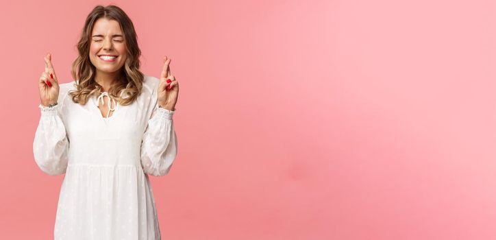 Portrait of hopeful lucky cute blond girl in white dress praying, anticiapte miracle, making wish to win, cross fingers good luck, smiling and close eyes and pleading god, pink background.