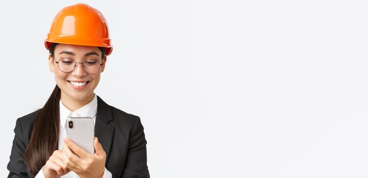 Close-up of smiling professional asian female industrial engineer, wearing safety helmet and business suit, using smartphone, looking pleased at mobile phone screen, standing white background.