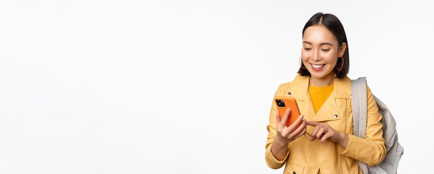 Image of happy girl traveller, tourist with backpack looking at smartphone, using mobile phone route app, standing over white background. Copy space