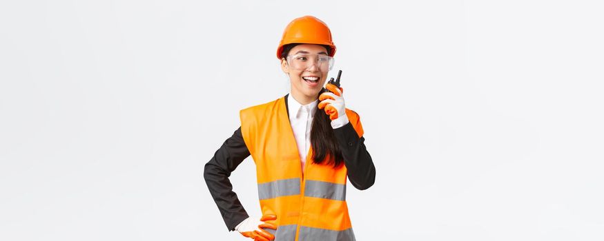 Confident pleased asian female engineer in safety helmet and uniform talking with chief architect using walkie-talkie. Satisfied construction technician contact team using radio phone.