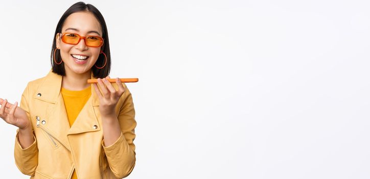 Image of happy asian girl talking on speakerphone, recording, translating her voice with mobile phone app, talking in smartphone dynamic, standing over white background.