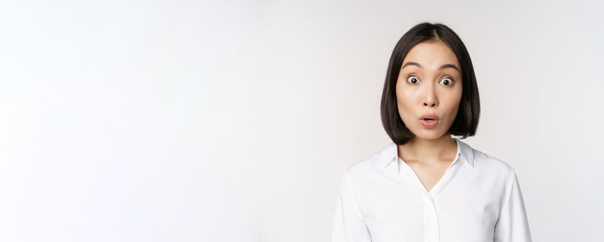 Close up portrait of young asian female model looking amazed at camera, smiling white teeth, standing against white background.