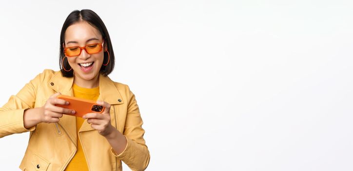 Stylish korean girl in sunglasses, playing mobile video game, laughing and smiling while using smartphone, standing over white background.