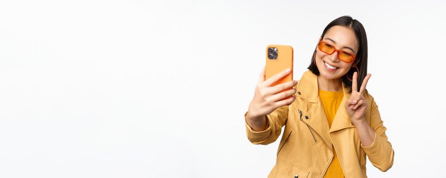 Stylish, beautiful asian girl in sunglasses, taking selfie on smartphone, posing for photo, holding mobile phone, white studio background.