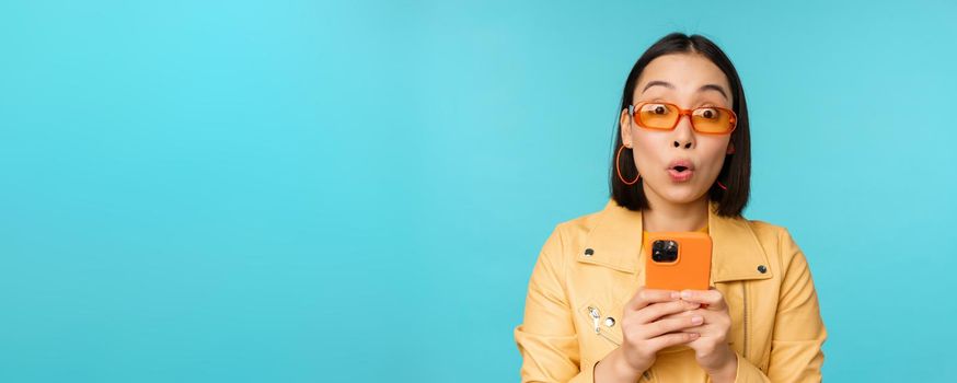 Image of asian girl in sunglasses, looking amazed and impressed, recording video or taking photo on smartphone, standing over blue background.