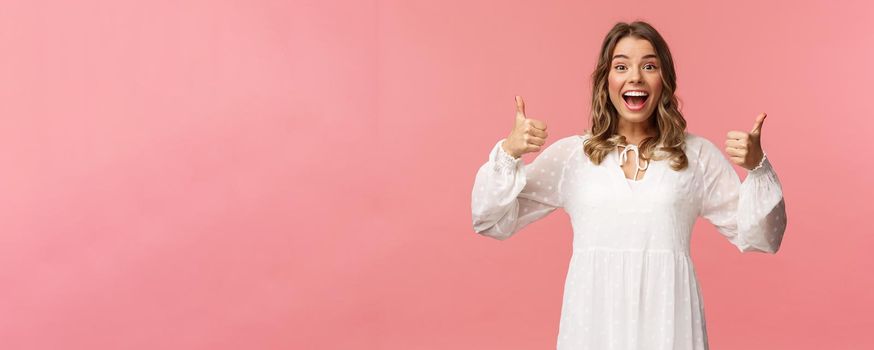 Portrait of excited feminine blond girl in white dress, show thumbs-up in approval, like being pleased, nod agreement, smiling fascinated, leave positive review, recommend product, pink background.