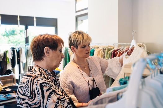 old woman with red hair, dressed in casual and modern way, mature woman blonde, dressed in long red skirt and pink jumper. two women in an exclusive fashion shop, buying clothes, mother and daughter enjoying a shopping day together. shopping concept. clothes seller in her small shop. natural light from window, sun rays, nature background plants, clothes racks, shop windows, clothes, Horizontal.