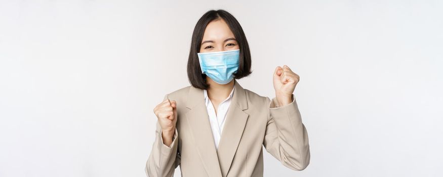 Coronavirus and business people concept. Happy businesswoman in medical face mask dancing, celebrating success, achievement, standing over white background.