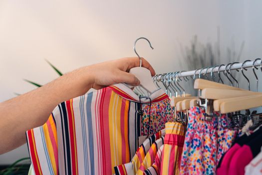 hand of a young woman taking a garment from a rack with clothes in fashion shop. women's clothes for sale. Shopping concept. dim light, soft and varied colours, fabric texture, white wooden clothes rack, summer and spring clothes.