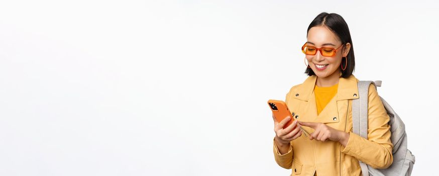 Stylish young asian woman tourist, traveller with backpack and smartphone smiling at camera, posing against white background. Copy space