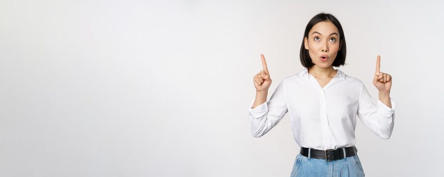 Enthusiastic asian business woman pointing, looking up with happy smiling face, showing company logo or banner, standing over white background.