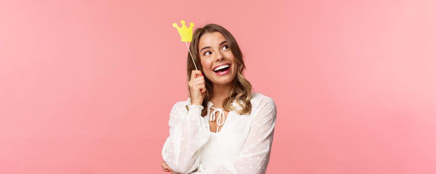 Spring, happiness and celebration concept. Close-up portrait of dreamy beautiful young blond girl imaging something cute and romantic, smiling look up daydreaming with crown on stick, pink background.