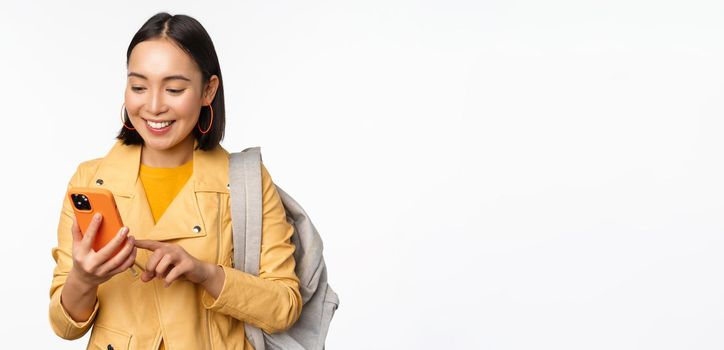 Image of happy girl traveller, tourist with backpack looking at smartphone, using mobile phone route app, standing over white background. Copy space