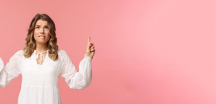 Skeptical and embarrassed blond caucasian woman in white cute dress, grimacing, cringe from seeing something disgusting, frowning doubtful and pointing fingers up strange thing, pink background.